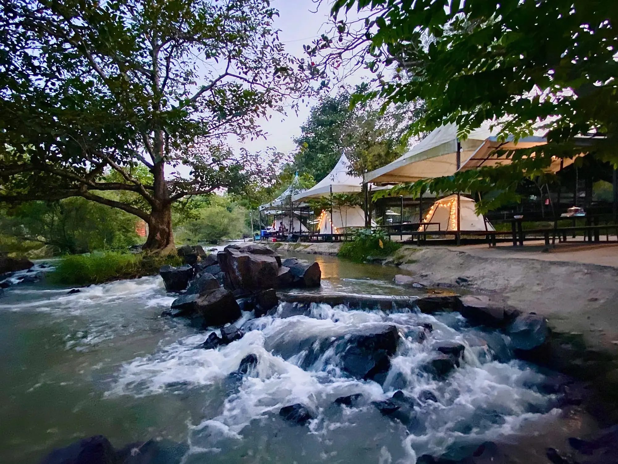 Ghềnh suối chảy xiết lúc chiều tối, bên bờ là dãy lều dưới mái che nhọn màu trắng sáng đèn